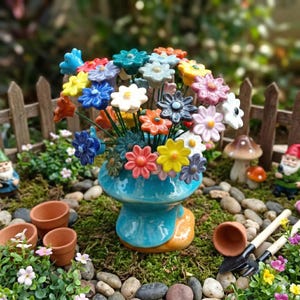 Op de afbeelding: Een keramisch bloemstuk in een blauwe vaas. De bloemen zijn in verschillende kleuren, waaronder blauw, geel, oranje en wit. De opstelling staat in een miniatuurtuin met kleine potten, een hek en kabouterbeeldjes.