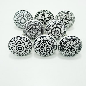 May include: Eight white ceramic cabinet knobs with detailed black mandala patterns. Each knob displays a different design, including floral and geometric shapes. The knobs are arranged in a semi-circle, highlighting their decorative appeal. The knobs are suitable for various furniture.