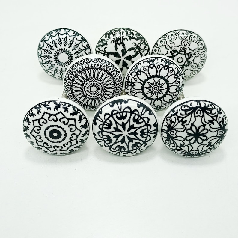 May include: Eight white ceramic cabinet knobs with detailed black mandala patterns. Each knob displays a different design, including floral and geometric shapes. The knobs are arranged in a semi-circle, highlighting their decorative appeal. The knobs are suitable for various furniture.