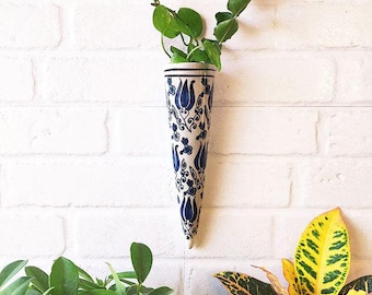 May include: A white ceramic wall vase with a blue floral pattern, holding green leafy plants. The vase is cone-shaped and hangs on a white brick wall. Other green plants are visible in the foreground.