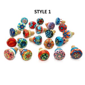 Peut inclure: Ensemble de bouchons de bouteille en céramique peints à la main. Chaque bouchon présente un design unique avec des couleurs et des motifs vifs, notamment des motifs floraux et géométriques. Le texte "STYLE 1" est visible.