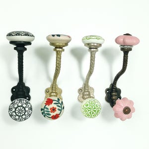 May include: Four decorative wall hooks with ceramic knobs. The hooks have different designs, including floral and geometric patterns, and a variety of colors such as black, white, pink, green, and gold. The hooks are made of metal.