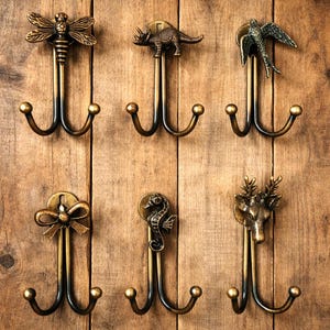 Peut inclure: Ensemble de six crochets muraux décoratifs en bronze. Chaque crochet présente un motif unique : libellule, dinosaure, oiseau, nœud, hippocampe et tête de cerf. Fixés sur une surface en bois, pour manteaux ou accessoires.
