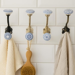 May include: Four decorative wall hooks with white and blue floral ceramic knobs. Two hooks have black metal stems, and two have gold-colored stems. A white towel and a wooden brush hang from the hooks.