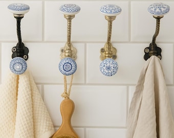 May include: Four decorative wall hooks with white and blue floral ceramic knobs. Two hooks have black metal stems, and two have gold-colored stems. A white towel and a wooden brush hang from the hooks.