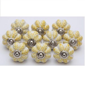 Peut inclure: Dix boutons de meuble en forme de fleur. Chaque bouton a une base blanche avec des motifs de feuilles jaunes et un centre en métal argenté. Ces boutons décoratifs sont conçus pour les meubles et la décoration intérieure.