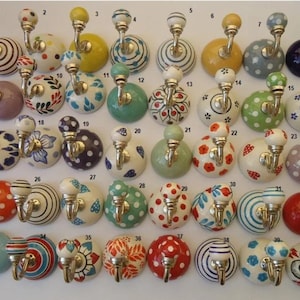 Könnte beinhalten: Eine Sammlung dekorativer Keramik-Wandhaken mit verschiedenen Designs. Die Haken weisen farbenfrohe Muster wie Punkte, Streifen und Blumenmotive auf. Jeder Haken hat einen Metallhaken und einen abgerundeten Keramikknauf.