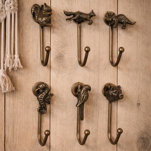 May include: A collection of six bronze-toned metal wall hooks, each with a different animal design. The hooks depict a squirrel, fox, rhinoceros, seahorse, parrot, and bear. They are mounted on a light brown wooden surface.