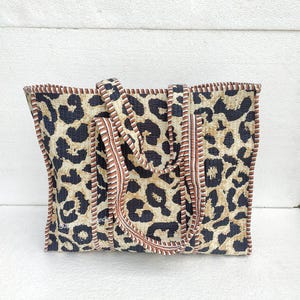 May include: A large tote bag with a leopard print design. The bag has brown and white stripes along the top edge and brown straps.
