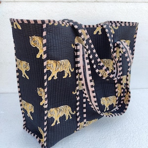 May include: A black quilted tote bag with pink and white striped handles. The bag features a repeating pattern of yellow and black tigers.