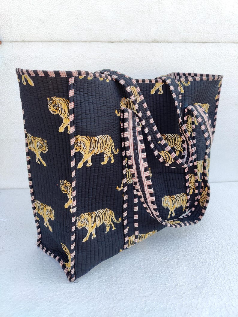 May include: A black quilted tote bag with pink and white striped handles. The bag features a repeating pattern of yellow and black tigers.