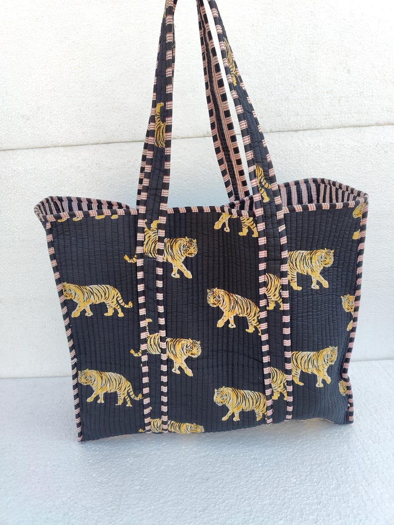 May include: A black quilted tote bag with pink and white striped handles. The bag features a repeating pattern of yellow tigers on a black background.