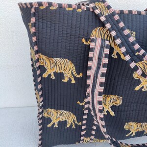 May include: A black quilted tote bag with pink and white striped handles. The bag features a repeating pattern of orange and black tigers.