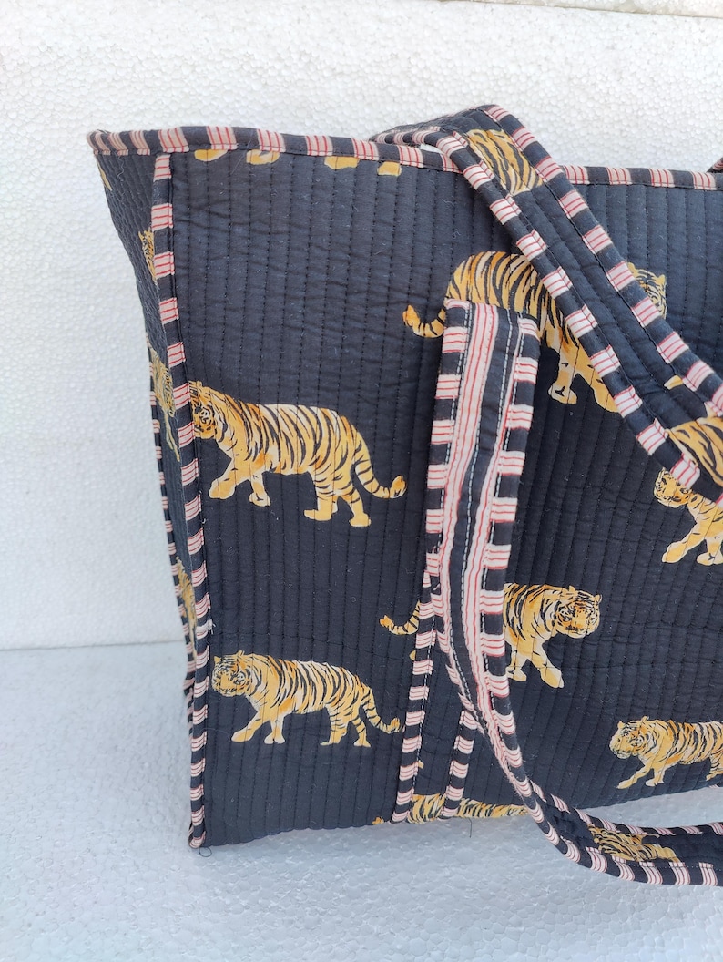 May include: A black quilted tote bag with pink and white striped handles. The bag features a repeating pattern of orange and black tigers.