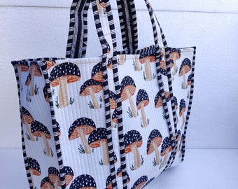 May include: A white tote bag with a black and white striped handle and a mushroom print. The bag is made of quilted fabric and has a black and white trim.