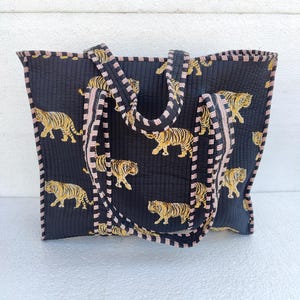 May include: A black quilted tote bag with pink and black striped trim. The bag features a repeating pattern of orange tigers on a black background.