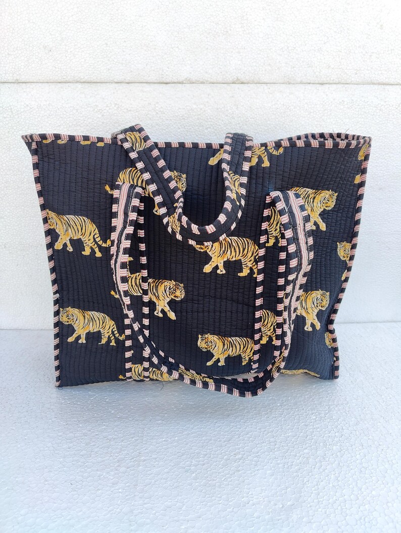 May include: A black quilted tote bag with pink and black striped trim. The bag features a repeating pattern of orange tigers on a black background.