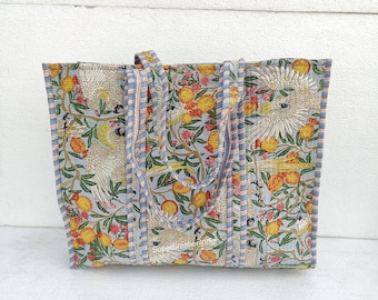 Op de afbeelding: Een grote tote bag met een blauwe achtergrond en een bloemenpatroon met gele citroenen, rode bloemen en witte vogels. De tas heeft blauw-wit gestreepte handvatten.