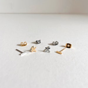 May include: A set of six silver and gold earrings. The earrings are small and simple, with a variety of shapes, including a cross, a bar, and a circle.