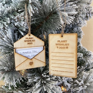 May include: Two wooden Christmas decorations shaped like envelopes with the text "Plant Wishlist 2021" on them. One ornament has a handwritten list inside, and the other has blank lines for writing.