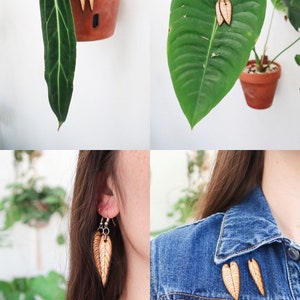 May include: A pair of wooden leaf earrings and a set of three wooden leaf pins. The earrings and pins are a light brown color with a wood grain pattern. The earrings are hanging from silver hooks. The pins are shown on a blue denim jacket.