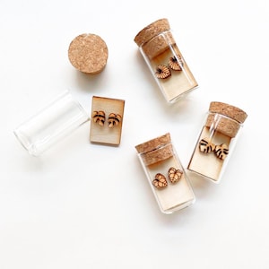 Op de afbeelding: Vier kleine glazen flesjes met kurken, elk met een paar houten oorbellen. De oorbellen zijn bladvormig en hebben een natuurlijke houten afwerking.