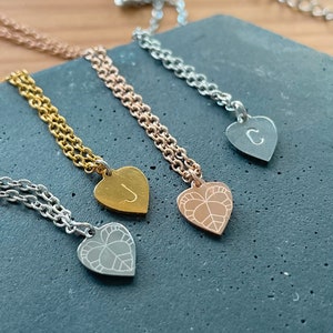 May include: Four silver heart necklaces with different charms. One necklace has a silver heart with a leaf design, one has a gold heart with the letter J, one has a rose gold heart with a leaf design, and one has a silver heart with the letter C.