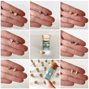 May include: A set of gold and silver earrings featuring a variety of leaf designs. The earrings are packaged in a small glass vial with a cork top. The vial has a label that reads "The Good Plant" and "Monstera Deliciosa".