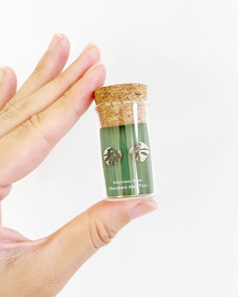 May include: A small glass vial with a cork top containing a pair of silver Monstera Albo Thai leaf earrings. The vial is labeled "Stainless Steel Monstera Albo Thai".