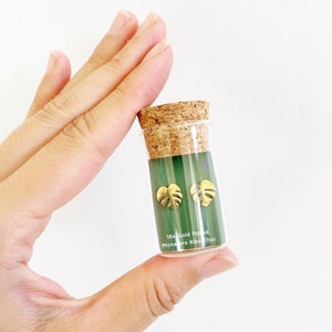 May include: A small glass vial with a cork top containing a pair of gold-plated Monstera Albo Thai leaf earrings. The vial has a green label with the text "18k Gold Plated Monstera Albo Thai".