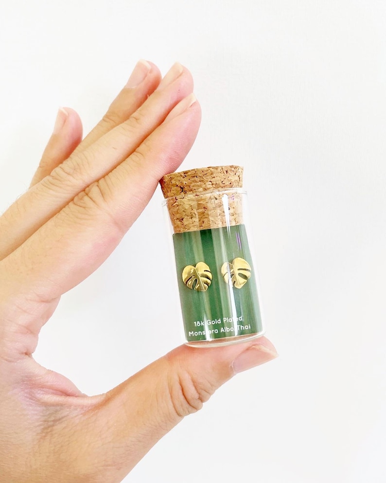 May include: A small glass vial with a cork top containing a pair of gold-plated Monstera Albo Thai leaf earrings. The vial has a green label with the text "18k Gold Plated Monstera Albo Thai".
