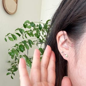 May include: A person's ear with multiple small gold earrings. The earrings are in the shape of a star, a small circle, and a small heart.