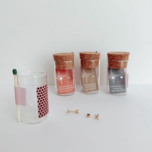 May include: Three small glass jars with cork tops containing gold star earrings. The jars are labeled "Flat Back Earrings" and have a pink, brown, and gray background. The earrings are displayed on a white surface with a matchstick and a glass jar.