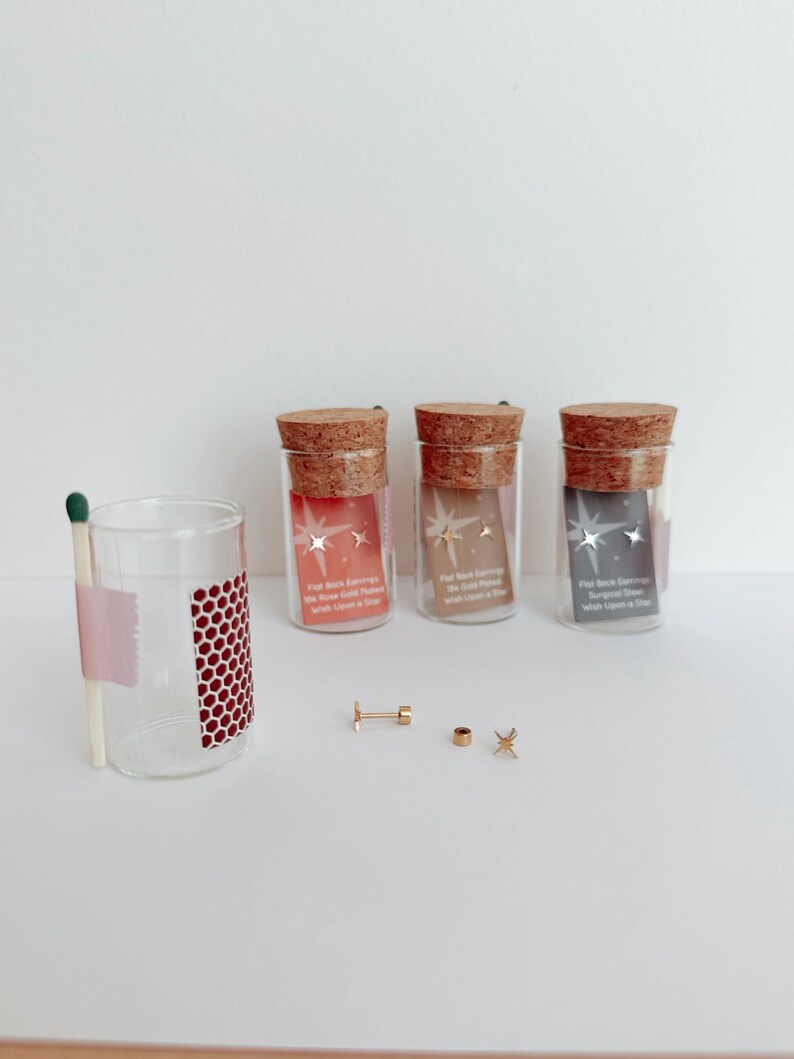 May include: Three small glass jars with cork tops containing gold star earrings. The jars are labeled "Flat Back Earrings" and have a pink, brown, and gray background. The earrings are displayed on a white surface with a matchstick and a glass jar.