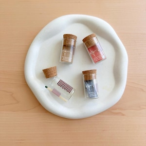 May include: Four small glass vials with cork tops, each containing a pair of gold star earrings. The vials are on a white ceramic plate. One vial contains a matchstick.