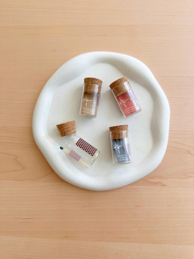 May include: Four small glass vials with cork tops, each containing a pair of gold star earrings. The vials are on a white ceramic plate. One vial contains a matchstick.