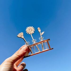 Puede incluir: Una exhibición de flores de madera en miniatura con tres diseños de flores diferentes. Cada flor se coloca en un pequeño vial de vidrio y se sujeta en un estante de madera. El fondo es un cielo azul claro y brillante.