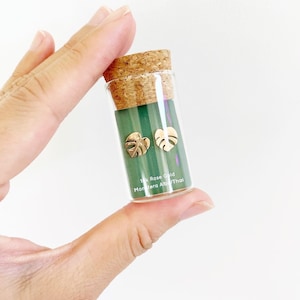May include: A small glass vial with a cork top containing a pair of gold Monstera Deliciosa leaf earrings. The vial has a green label that reads "14k Rose Gold Monstera Albo/Thai".