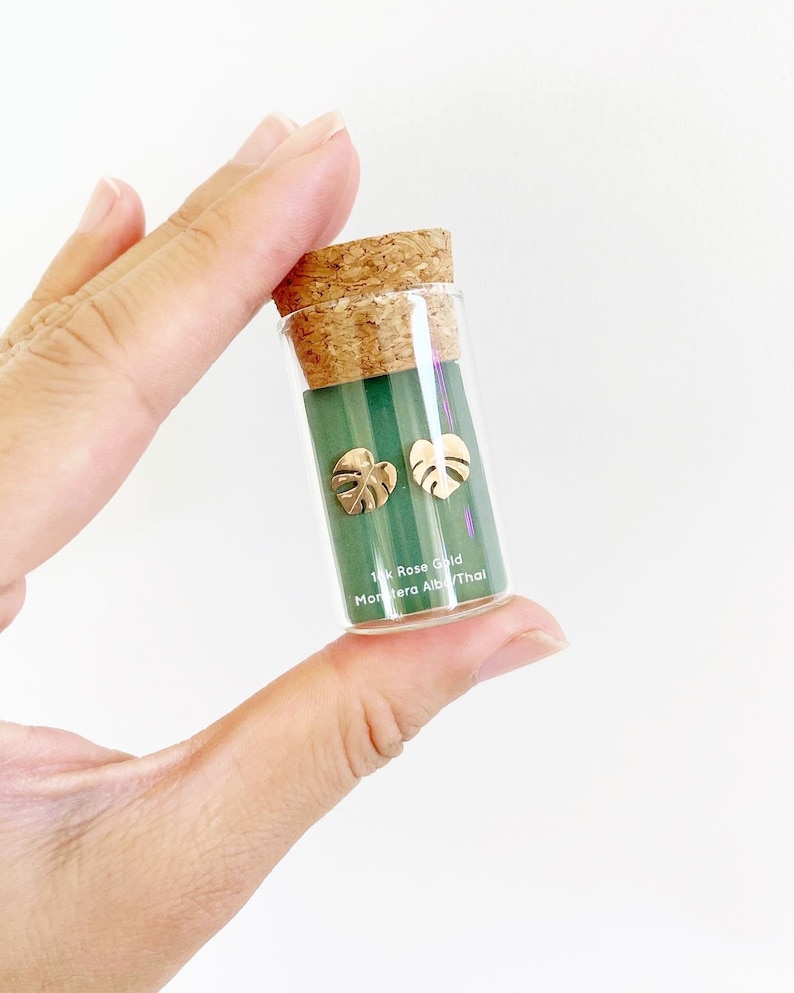 May include: A small glass vial with a cork top containing a pair of gold Monstera Deliciosa leaf earrings. The vial has a green label that reads "14k Rose Gold Monstera Albo/Thai".