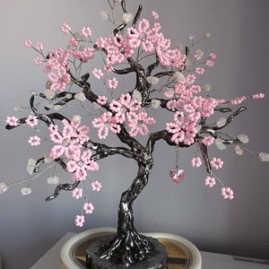 May include: A pink beaded wire tree with a crystal heart hanging from a branch. The tree is on a white heart-shaped base.
