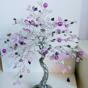 Puede incluir: Una escultura de árbol de alambre plateado decorada con cuentas y cristales morados y rosados. El árbol tiene un tronco retorcido y una base de piedra.