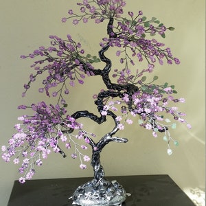 Może przedstawiać: Drzewko bonsai z fioletowych koralików z czarną ramą z drutu i srebrną podstawą. Drzewko stoi na czarnej powierzchni.