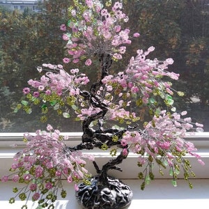May include: A beaded bonsai tree with pink and green beads. The tree is on a black base and is sitting on a white surface.