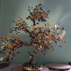 May include: A handcrafted wire tree sculpture with a bronze-colored base and trunk. The tree's branches are adorned with amber, gold, and clear beads, creating a decorative bonsai-like appearance. The tree sits on a wooden slice base.