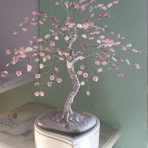 Op de afbeelding: Een zilveren draadboom met roze kralen en heldere kristallen, die op een wit hartvormig muziekdoosje staat.