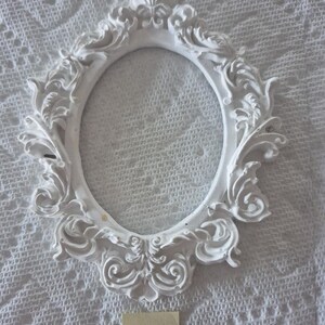 May include: A white, ornate, oval picture frame with intricate scrollwork and floral details. The frame is empty, with a smooth, blank interior. The frame is made of a solid material and is set against a textured white background.