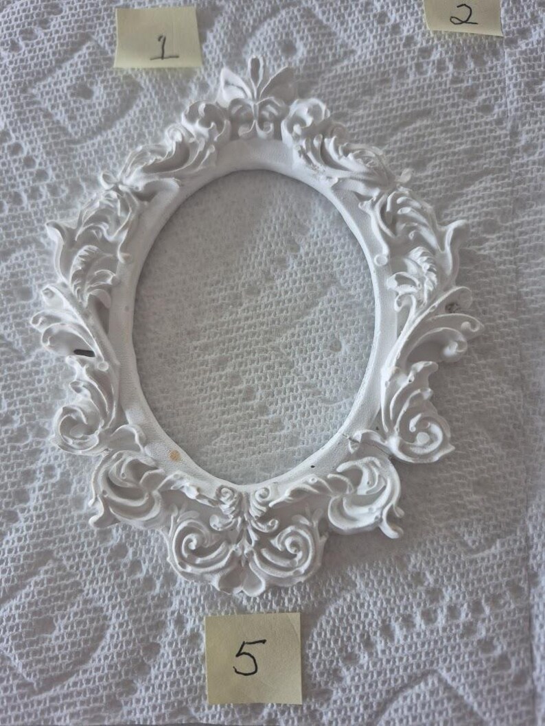 May include: A white, ornate, oval picture frame with intricate scrollwork and floral details. The frame is empty, with a smooth, blank interior. The frame is made of a solid material and is set against a textured white background.
