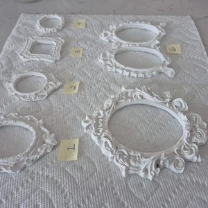May include: A collection of white, ornate picture frames in various shapes and sizes, including oval, round, and square designs. The frames are arranged on a white textured surface, with small yellow numbered tags next to each frame.