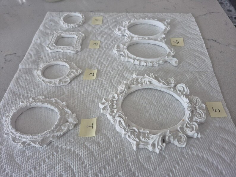 May include: A collection of white, ornate picture frames in various shapes and sizes, including oval, round, and square designs. The frames are arranged on a white textured surface, with small yellow numbered tags next to each frame.