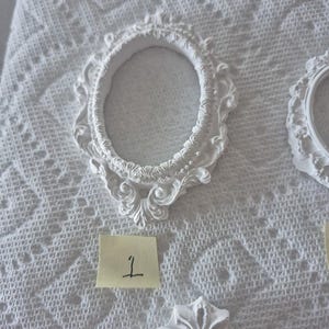 May include: White, ornate oval picture frame with decorative scrollwork. The frame is made of a solid material and is placed on a white, textured surface. A small yellow sticky note with the number "1" is visible.
